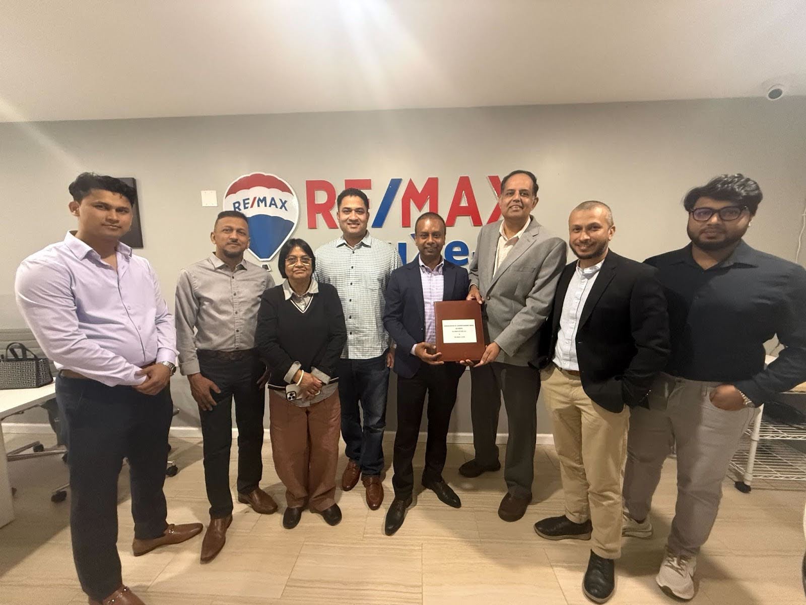 A group of eight people stands together indoors, after recognition of partnership, in front of a RemaxLuxe logo.